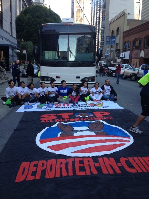 Photo of activists sitting in front of a bus with a banner reading 