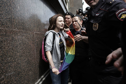 Photo of a person holding a rainbow flag being pushed against a wall by police officers [Photo credit: RT.com]