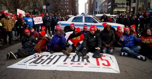 Photo of a group of people sitting in the middle of a street with a Fight for 15 banner in front of them
