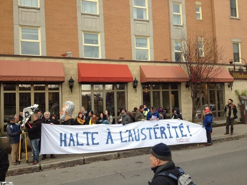 Photo : devant un bâtiment de briques rouges, une vingtaine de personnes tiennent une banderole Halte à l'austérité. Marionnette géante de Couillard.