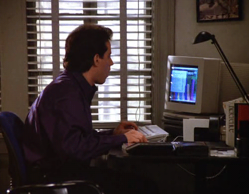Jerry Seinfeld sits at desk looking at computer.
